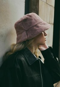 Chapeau seau en fausse fourrure rose avec un large bord et une surface texturée, porté avec une veste en cuir noire, positionné contre un mur neutre.