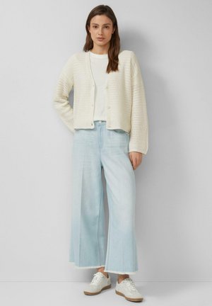 Kobieta w kremowym swetrze z dzianiny, białym t-shircie, jasnoniebieskich szerokich jeansach i białych tenisówkach stoi na tle jednolitego tła.