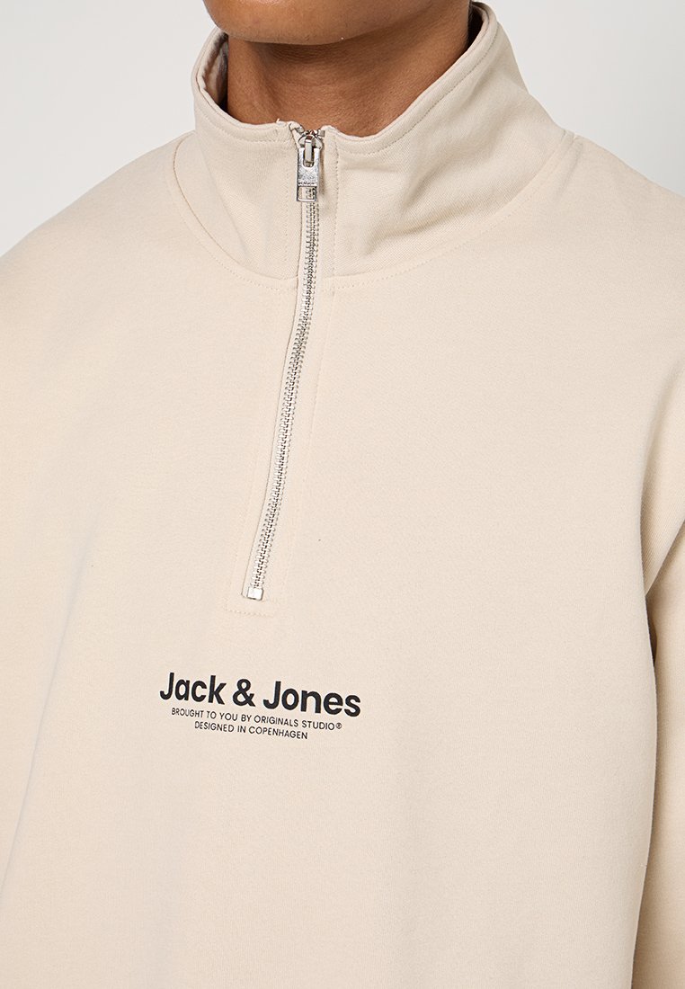 Sudadera con cremallera en color beige, con cuello alto y fabricada en un tejido suave. Presenta texto de logotipo negro en la parte delantera, detalle de cremallera plateada y un ajuste relajado.