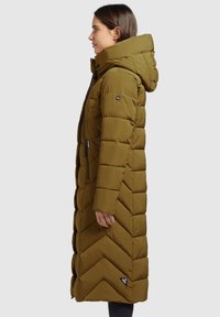 Maslinasto zelena puffer jakna s kapuljačom, koja ima žicasti uzorak, bočne džepove sa patentnim zatvaračem i oznaku logotipa blizu došivene ivice. Dužina do sredine butina.