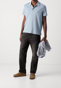 Ljusblå pikétröja med ett texturerat mönster, kombinerad med mörka jeans. Modellen håller i en vikt randig skjorta och bär bruna mockaklogs.