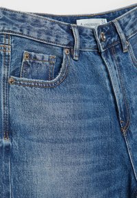 Jean en denim bleu avec bouton en métal, passants pour ceinture, poche avant et étiquette de taille visible indiquant 34.