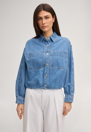 Kobieta o prostych brązowych włosach, ubrana w zapinaną na guziki niebieską dżinsową kurtkę z dużymi przednimi kieszeniami i luźne białe spodnie na jednolitym tle.