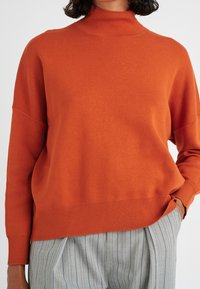 Kvinna iklädd en löst sittande orange tröja med neddragna axlar och grå randiga byxor, med ena handen i fickan.