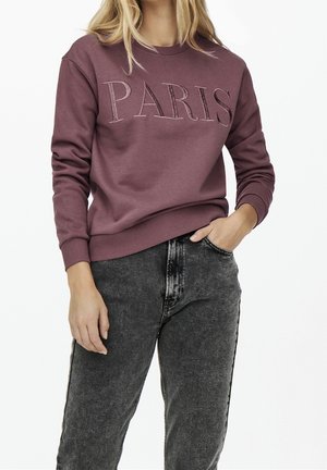 Femme portant un sweat mauve avec le texte "PARIS" et un jean noir effacé par acide, une main dans la poche, debout devant un fond blanc.