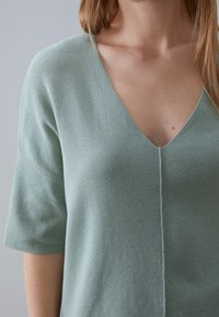 Maglione a V verde chiaro, lavorato a maglia, con maniche corte e un dettaglio cucito centrale. Tessuto morbido e vestibilità comoda.