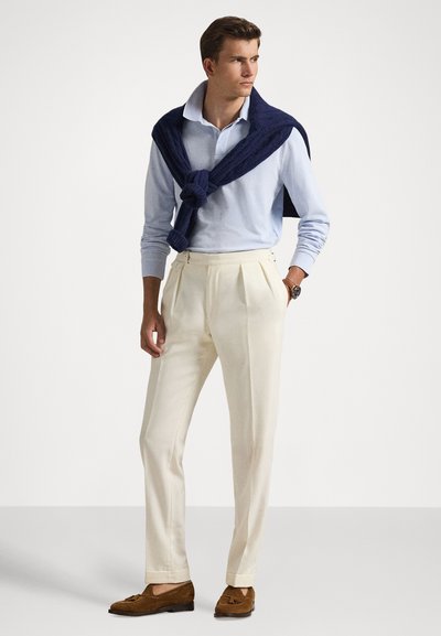 Νεαρός άνδρας με ανοιχτό μπλε μακρυμάνικη πόλο, σκούρο μπλε πουλόβερ περασμένο στους ώμους, κρεμ πτυχωτό παντελόνι, καφέ μοκασίνια και ρολόι χειρός, όρθιος.