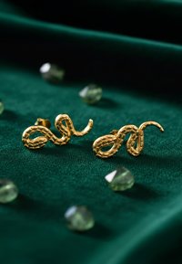 Orecchini a forma di serpente in oro con dettagli testurizzati, esposti su un tessuto verde, circondati da piccole gemme verdi.