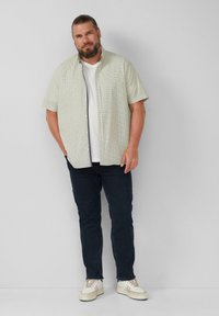 Koszula z krótkim rękawem, zapinana na guziki, w jasnym zielonym kolorze z wzorem siatki, połączona z ciemnymi dżinsami i beżowymi sneakersami. Luźny, casualowy krój.