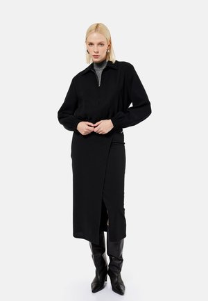 Blondynka w czarnej midi spódnicy z rozcięciem z przodu, czarnej kurtce na szarym golfie i czarnych wysokich botkach, stojąca na białym tle.