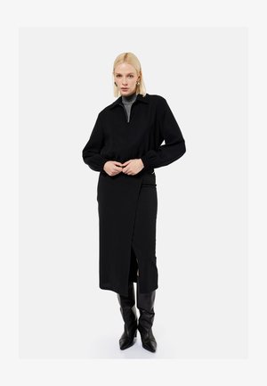 Blondynka w czarnej midi spódnicy z rozcięciem z przodu, czarnej kurtce na szarym golfie i czarnych wysokich botkach, stojąca na białym tle.