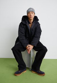 Giubbotto puffa nero con cappuccio, camicia marrone sotto, pantaloni neri, scarpe blu e un berretto lavorato a maglia grigio, seduto su una valigia argentata.