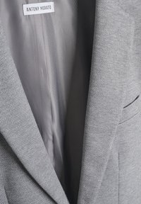 Sivi sako s uzorkom pod nazivom houndstooth, sa glatkim unutarnjim podstava. Oznaka kaže "ANTONY MORATO." Istaknuti detalji revera i džepova.