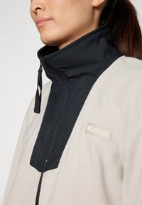 Fleece bunda s vysokým čiernym golierom a krémovou telom. Výrazné logo Columbia na ľavej strane hrude a zips s detailom na ťahadle.
