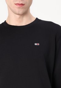 Svart bomullslångärmad tröja med rund halsringning, med en liten broderad logotyp i vitt, rött och blått på bröstet.