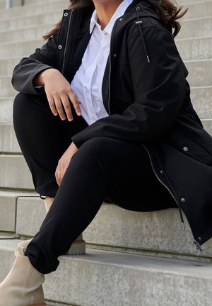 Giacca nera impermeabile, camicia bianca con colletto, leggings neri e stivaletti beige. Tessuto strutturato. Seduta su gradini di pietra.