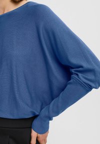 Pull bleu en tissu tricot doux, doté d'une coupe décontractée, de larges manches et d'une texture côtelée au niveau des poignets. Le design est minimaliste.