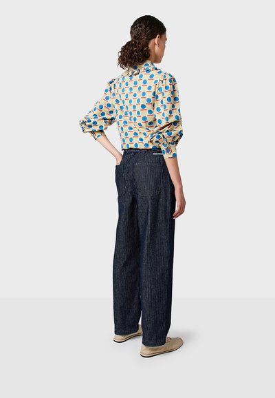 Bunte Bluse mit Polka-Dots und Puffärmeln, kombiniert mit navyblauen, gestreiften, weiten Hosen und beigen Slippern, von hinten betrachtet.
