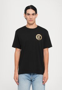 Svart bomulls T-skjorte med korte ermer, med et sirkulært gulllogo på venstre bryst. Kombinert med lyseblå jeans.