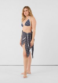 Bikini blu e bianco a motivi con spalline a collare, abbinato a un sarong regolabile coordinato. Modello indossa ciabatte bianche.