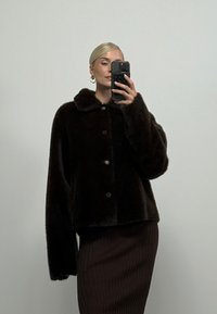 Femme blonde aux cheveux plaqués en arrière porte un manteau en fourrure noir et une jupe foncée côtelée, prenant un selfie dans un miroir avec un smartphone noir.