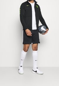 Chaqueta negra con cremalleras y detalles verdes, camiseta blanca, pantalones cortos negros con ribetes blancos, calcetines blancos y zapatillas negras y blancas, sosteniendo un balón de fútbol.