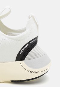 Gros plan sur le talon d'une chaussure de course blanche Nike avec tissu Flyknit et texte "Nike Free" sur la semelle texturée en noir et beige.