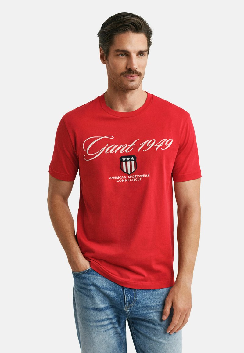 Rotes Baumwoll-T-Shirt mit weißem "Gant 1949"-Text und einem marineblauen Wappenlogo mit drei Sternen. Kurze Arme und Rundhalsausschnitt.
