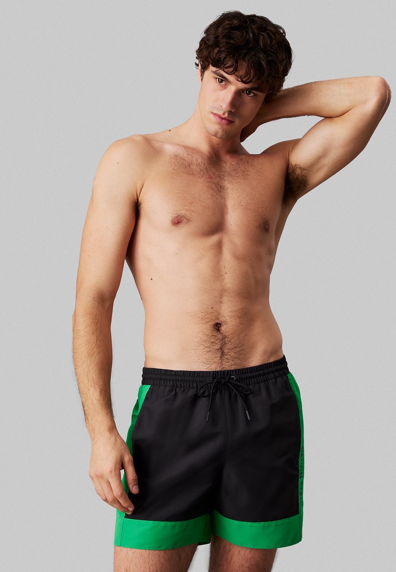 Czarne spodenki kąpielowe z zielonymi bocznymi panelami, elastyczną talią i sznurkiem do regulacji. Gładka tekstura, długość do połowy uda, z kontrastującymi akcentami kolorystycznymi.