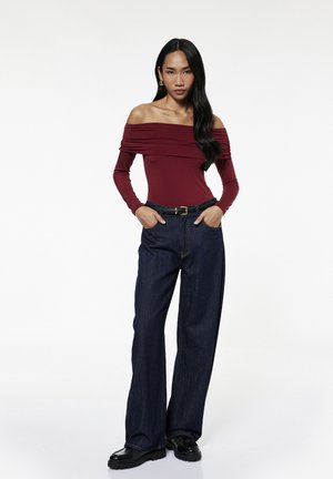 Kobieta stoi z rękami w kieszeniach, nosi bordowy top odkrywający ramiona, ciemne szerokie dżinsy z paskiem, czarne buty i złote kolczyki koła.
