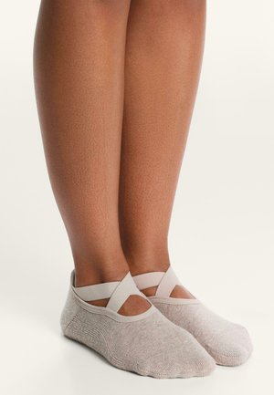 Gambe e piedi inferiori che indossano calzini stile balletto grigio chiaro con fasce elastiche incrociate su uno sfondo bianco uniforme.