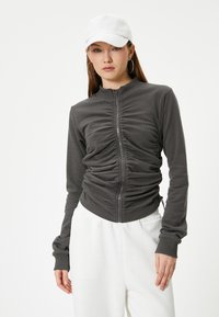 Grijze zip-up jas met gerimpelde details, lange mouwen en een getailleerd silhouet, gecombineerd met een witte pet en witte broek. Soepele textuur.
