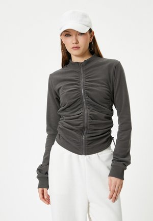 Veste zippée grise avec des détails plissés, manches longues et silhouette ajustée, associée à une casquette blanche et un pantalon blanc. Texture lisse.