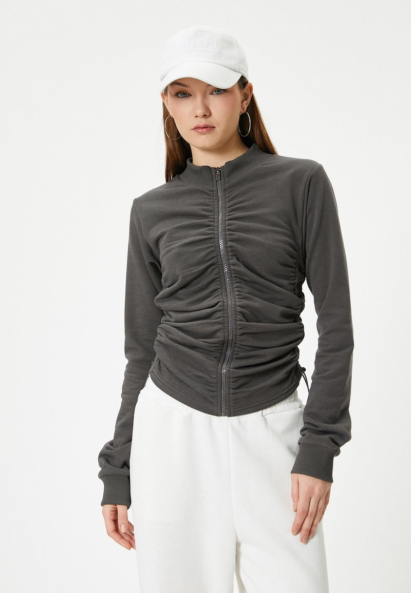 Grijze zip-up jas met gerimpelde details, lange mouwen en een getailleerd silhouet, gecombineerd met een witte pet en witte broek. Soepele textuur.