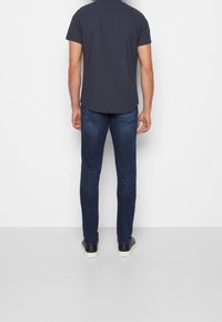 Camicetta a maniche corte blu navy con motivo texturizzato, abbinata a jeans slim-fit blu scuro e sneaker bianco e nero. Vista da dietro.