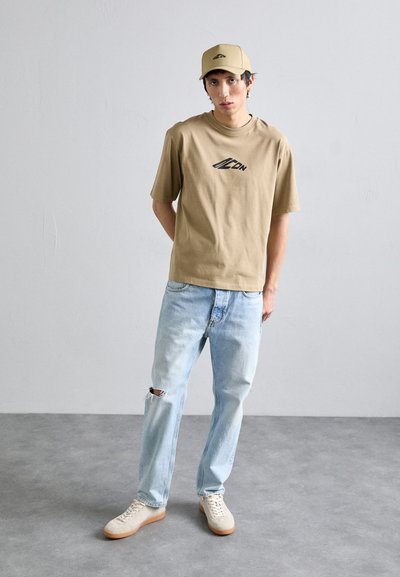 Tan bomulds T-shirt med logo, lyseblå jeans med hul ved kn�æet, beige sneakers og matchende kasket. Minimalistisk design mod en neutral baggrund.