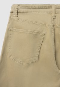 Pantalon en coton beige vu de dos, présentant une poche carrée avec un rabat triangulaire, des coutures surpiquées et une passants de ceinture à la taille.