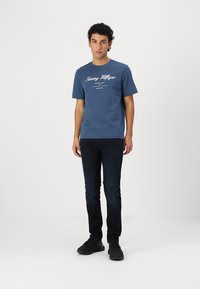 Kék pamut póló fehér "Tommy Hilfiger" felirattal és díszítőelemekkel, sötét farmernadrággal és fekete sportcipővel párosítva.
