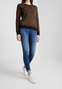 Een bruine luipaardprint trui met zwarte randen, gecombineerd met blauwe skinny jeans en witte sneakers. Zachte textuur, casual stijl.