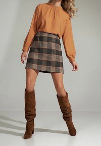 Blusa marrone a maniche lunghe con collo arricciato, abbinata a una gonna corta a quadri nei toni del nero, beige e arancione, e stivali al ginocchio in suede marrone.