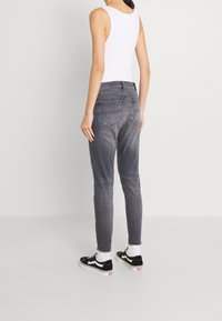 Mujer con jeans grises deslavados, camiseta sin mangas blanca, calcetines blancos y zapatillas negras de corte bajo, de pie contra un fondo claro y sencillo.