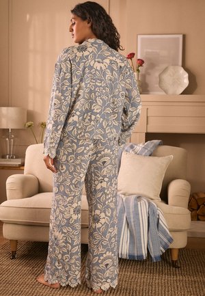 Woman standing barefoot in blue and white floral pajama set in cozy living room with beige sofa, striped throw, and flowers on mantel.