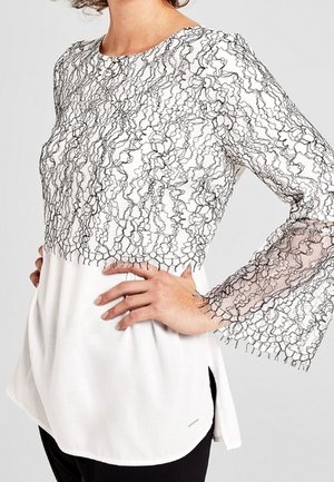 Biała bluzka z czarną koronkową nakładką, długie rozkloszowane rękawy, gładka tkanina pod koronką, zakrzywiony dół i boczne rozcięcia dla swobody ruchów.