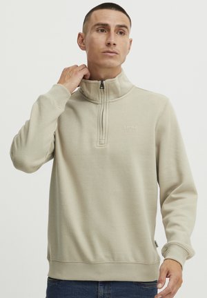 Beżowy sweatshirt z zamkiem na szyi, wykonany z miękkiego materiału, z wysokim kołnierzem, ściągaczami przy rękawach oraz małym logo na piersi.