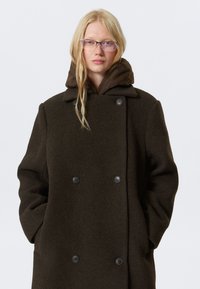 Double-breasted coat in dark green wool, featuring a hooded brown inner layer, large black buttons, and side pockets.