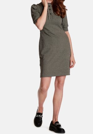 Vrouw draagt een knie-lange, olijfgroene jurk met korte mouwen en knopen op de borst, gecombineerd met zwarte loafers met zilveren kettingen.