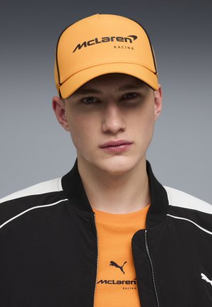 Junger Mann mit einer orangefarbenen McLaren Racing Mütze, passendem orangefarbenen McLaren Hemd und schwarzer Puma-Jacke mit weißen Streifen vor einem grauen Hintergrund.