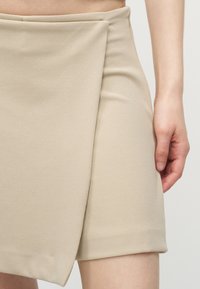 Gros plan sur une jupe beige de style portefeuille portée par une personne, montrant le détail du tissu plié et sa main droite reposant à côté de la jupe.