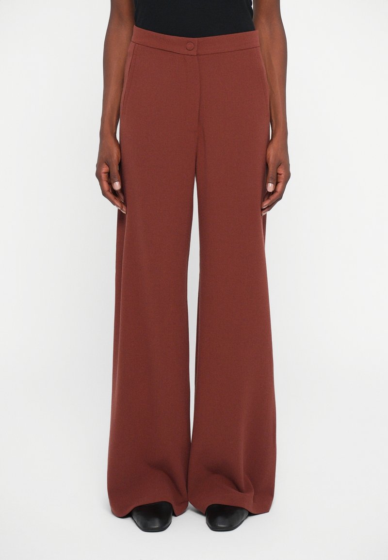 Personne portant un pantalon large taille haute couleur rouille avec une ceinture boutonnée et des chaussures noires à bout carré.