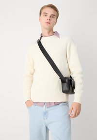 Suéter de canalé crema sobre una camisa a rayas burdeos y blanca, con unos vaqueros azul claro. Lleva un pequeño bolso cruzado negro.
